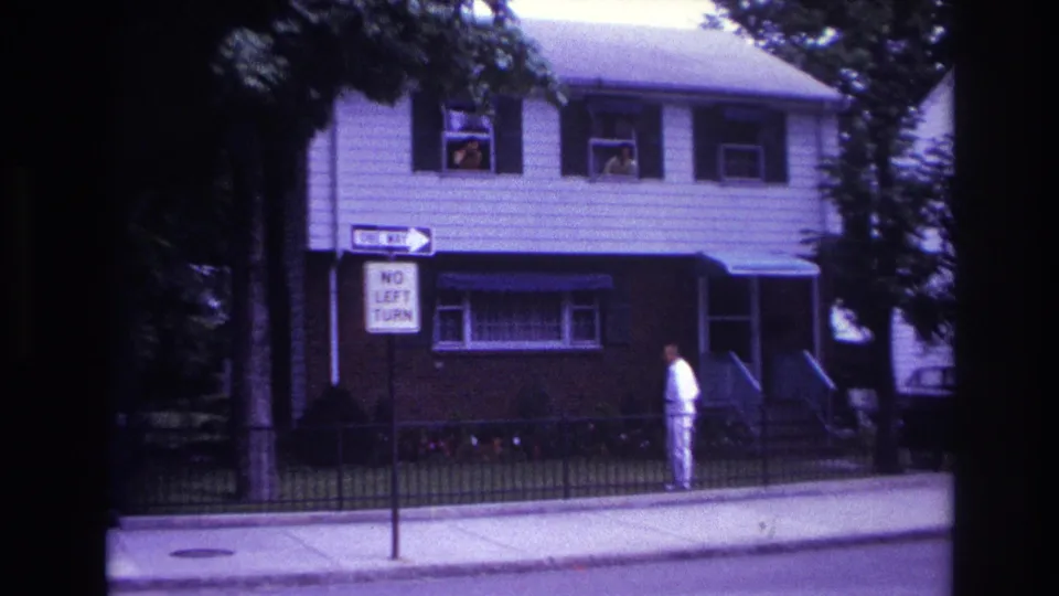 1967 Rockaway Beach residential street frame with a man in white beside a fenced yard and traffic signs in front of a two-story house.