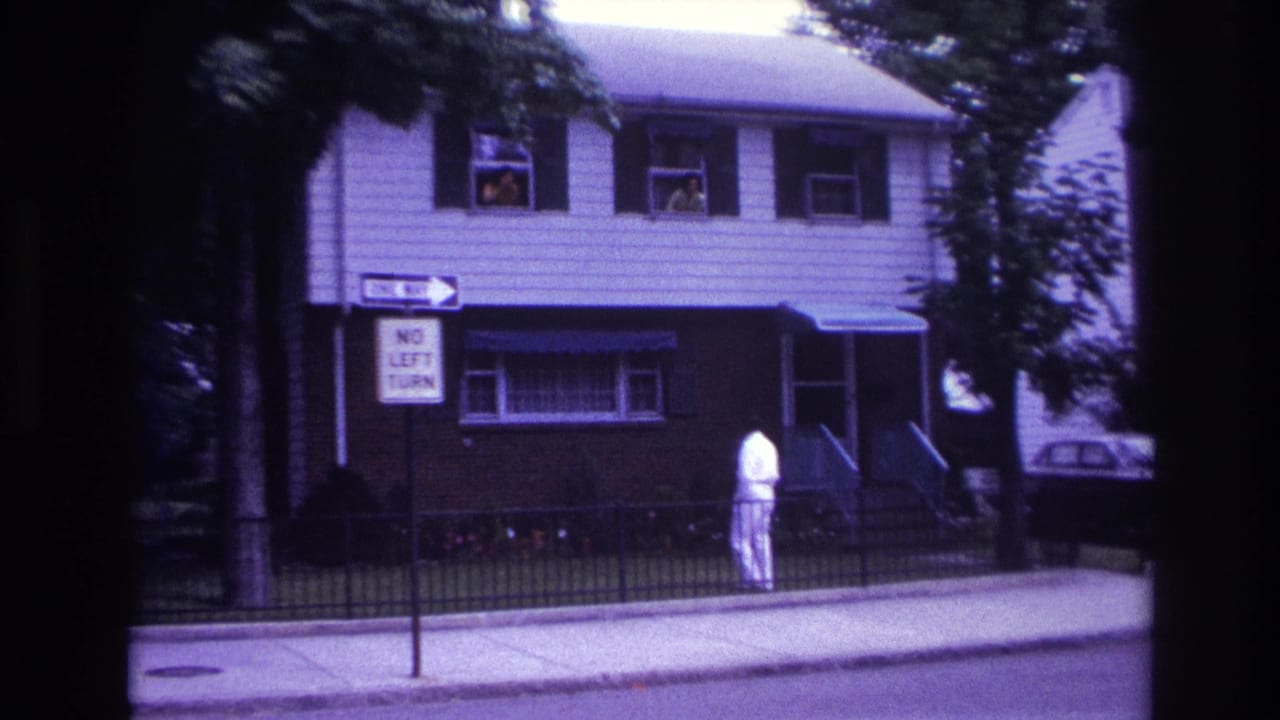 Man in a white suit stands on a sidewalk outside a two-story house with people in upstairs windows.