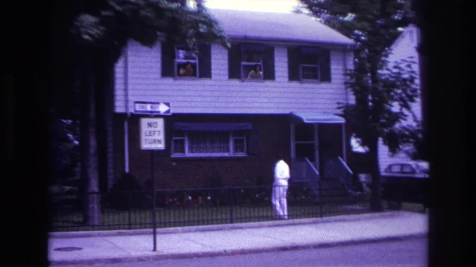 1967 Rockaway Beach home-movie frame showing a man in white near a fenced house, with two people visible in upper windows.