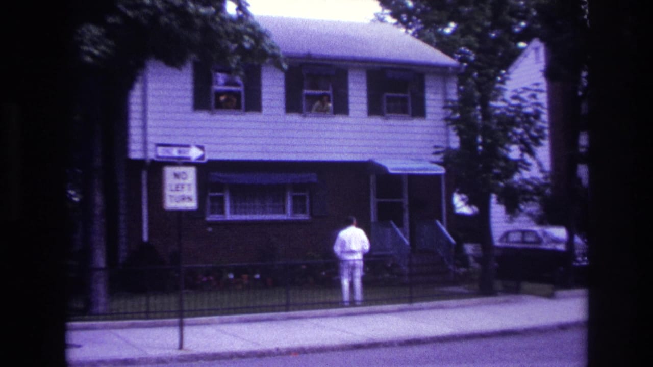 Man in white stands near the front fence of a house while two people remain visible in upstairs windows.