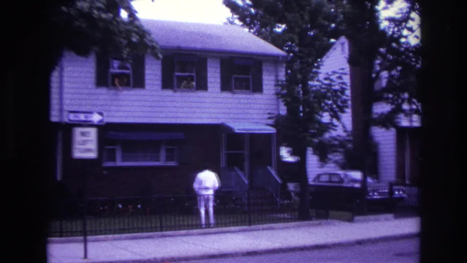 Rockaway Beach 1967 home-movie still showing a man in white on the sidewalk before a two-story house with people at windows.