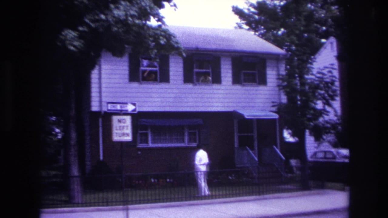 Man in white walks past a two-story house as people appear in upstairs windows.