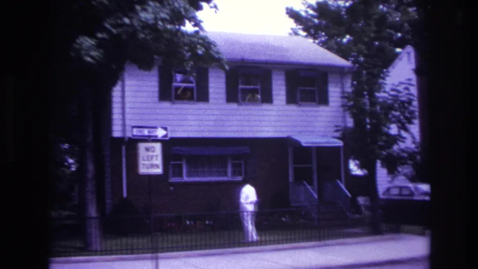 1967 Rockaway Beach home-movie frame showing a man in white near a house with people watching from upper windows.