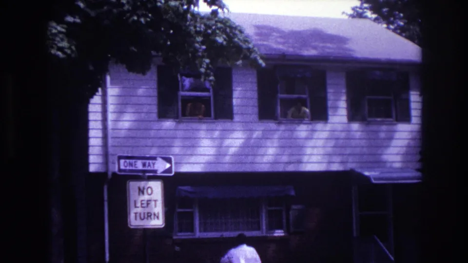 1967 home-movie view of a Rockaway Beach house facade with people at windows and traffic signs in front.