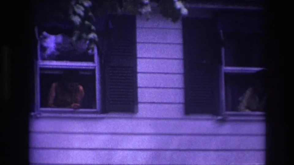 Rockaway Beach 1967 home-movie frame showing a woman at the left upper window and a light-clothed figure near the right window on a shuttered facade.