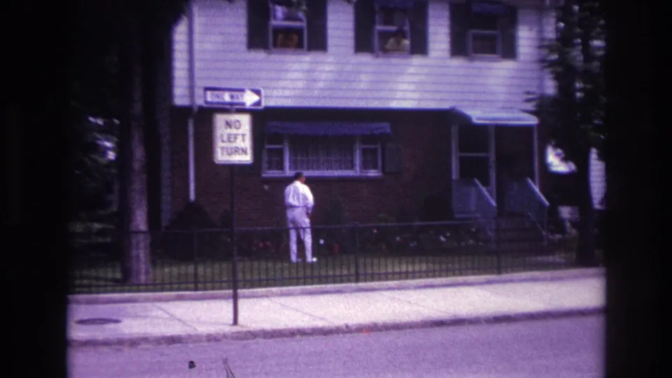 Wider 1967 Rockaway Beach home-movie view of a residential house, sidewalk signs, and a man in light clothing.