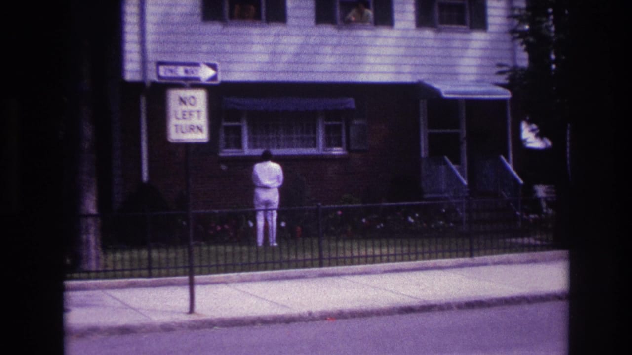 Man in white stands with his back to the camera in front of a house and traffic signs.