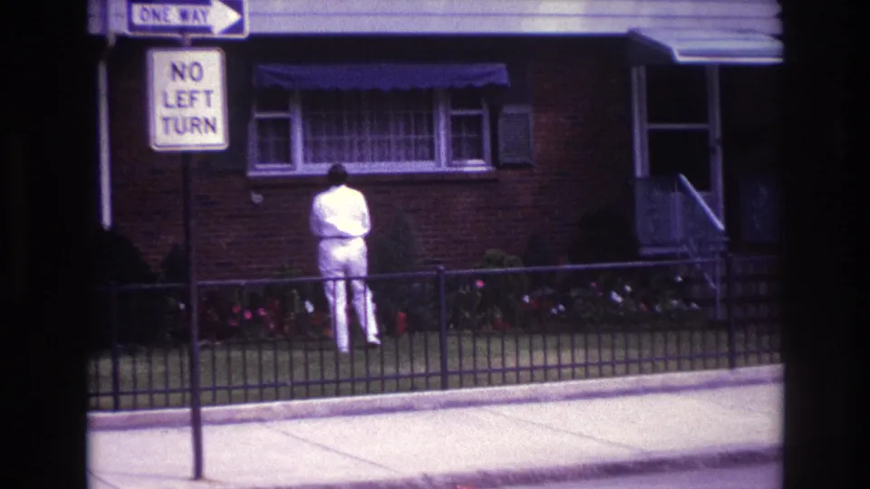 Closer 8mm frame of a man in white facing the front of a Rockaway Beach house, with fence, flowers, and street signs nearby.