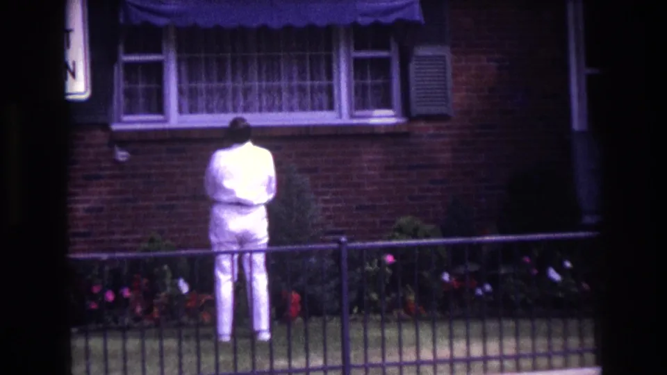 Tight 1967 home-movie view of the white-suited figure from behind, framed by a fence, flowers, brick wall, and curtained window.