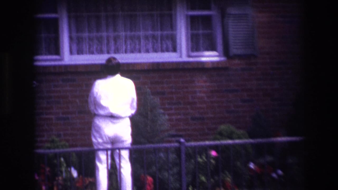 In Rockaway Beach home-movie footage, a man in a white suit stands with his back to the camera before a brick house window.
