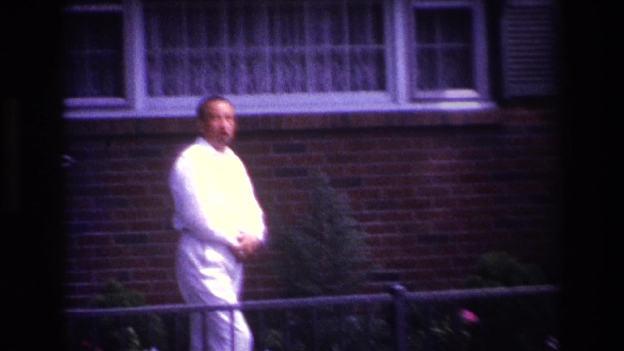 Man in a white suit turns toward the camera in front of a brick house.