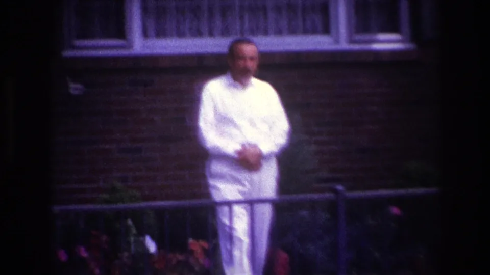 A softly focused 8mm frame shows the man in a white suit standing behind a fence with flowers and a brick house behind him.