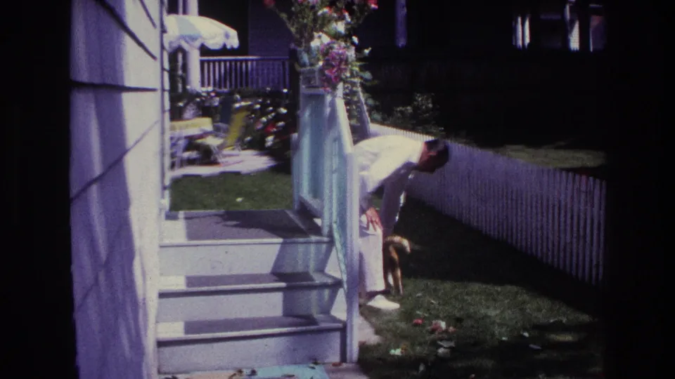 In the backyard, the man in a white suit bends near porch steps, flowers, lawn, a white picket fence, and a small dark animal.