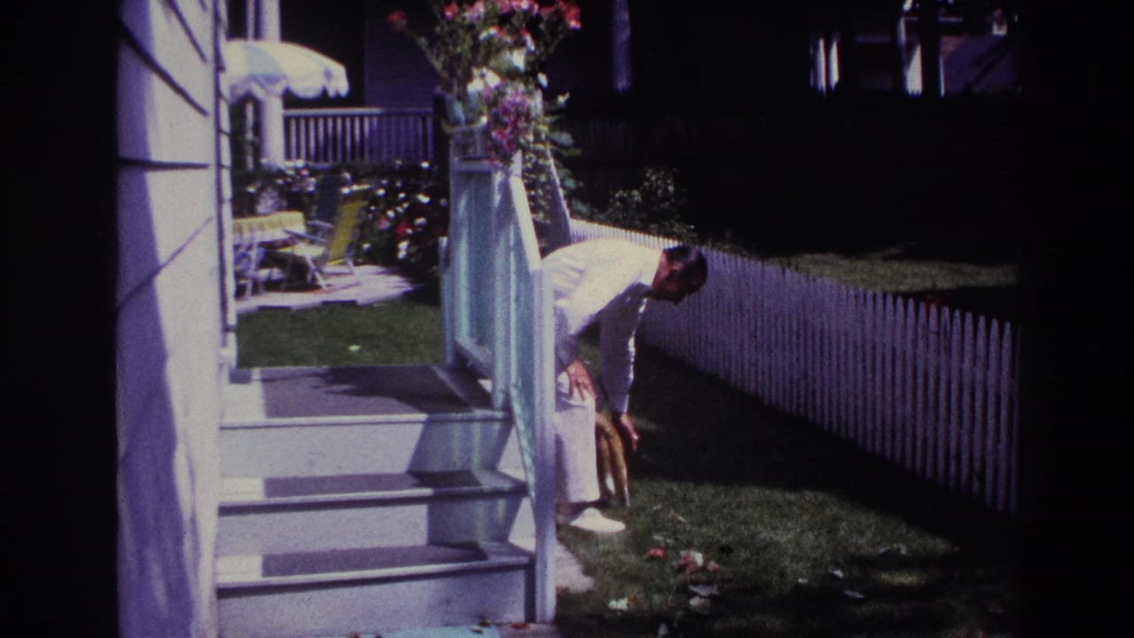 1967 Rockaway Beach home-movie frame showing a woman bending toward a cat beside porch steps, lawn, flowers, and a white picket fence.