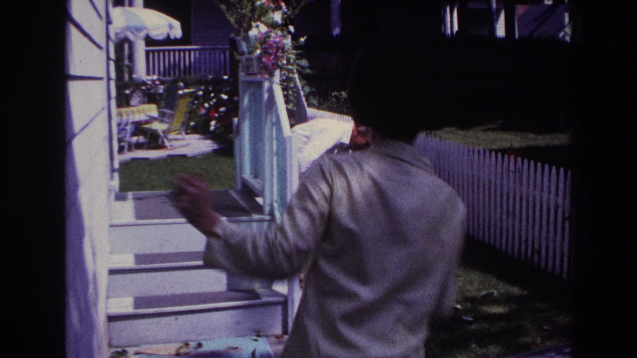 Man in a light jacket walks into the yard near porch steps and a white fence.