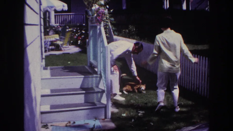 1967 Rockaway Beach home-movie frame showing a man approaching while a woman bends toward a ginger cat near porch steps and a fence.