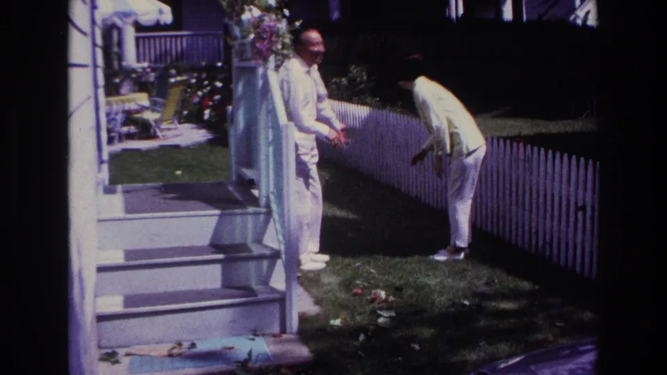 1967 Rockaway Beach frame showing two adults by a white picket fence and porch steps, with one bending toward the grass where the cat had been.
