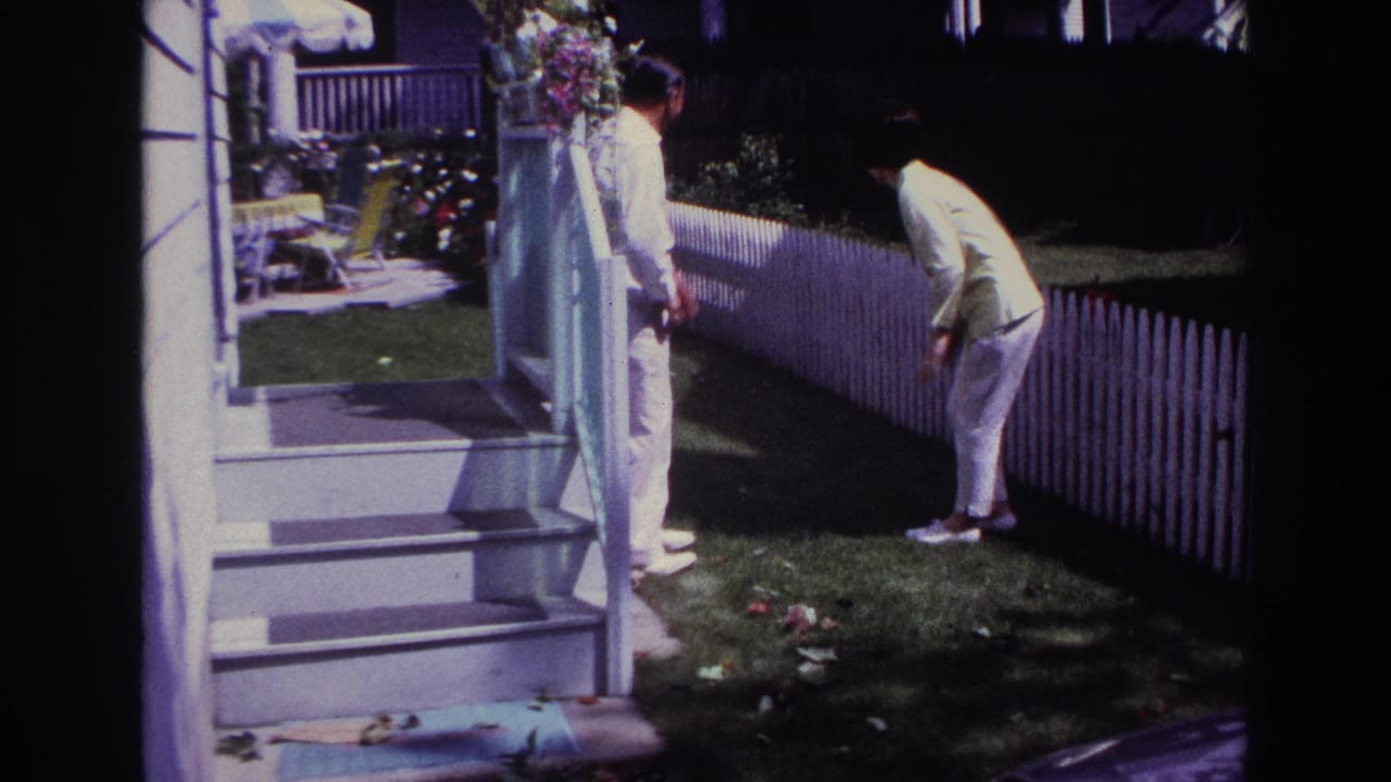 Man on porch steps faces a woman bending near a white picket fence in a sunny garden.