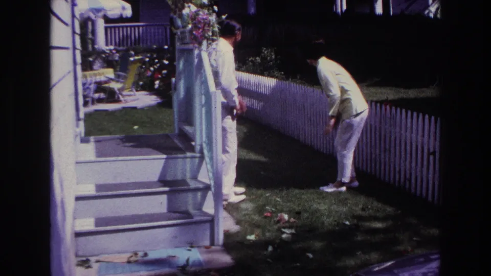 1967 Rockaway Beach home-movie frame showing a man on porch steps and a woman bending beside a white picket fence in a lawn garden.