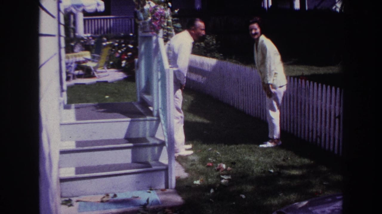 1967 Rockaway Beach backyard frame with a man and woman facing each other near porch steps, lawn, and a white picket fence.