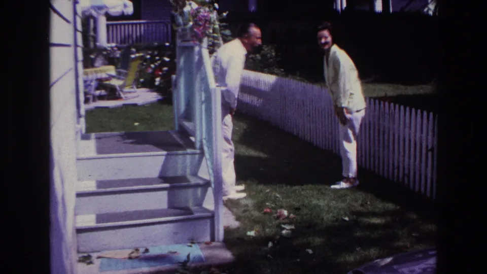 1967 Rockaway Beach backyard frame with a man and woman facing each other near porch steps, lawn, and a white picket fence.