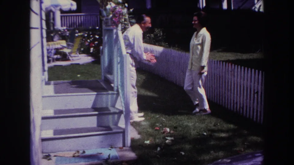 1967 Rockaway Beach home-movie frame showing a man gesturing to a woman in a light outfit near porch steps and a picket fence.