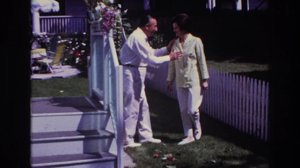 1967 Rockaway Beach backyard frame showing a man with his arm around a woman near porch steps, lawn, and a white picket fence.