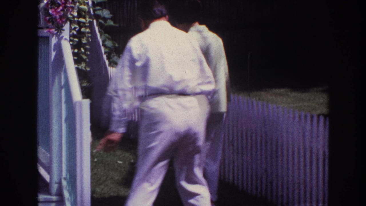 Two adults walk past a white fence near garden steps in bright sunlight.