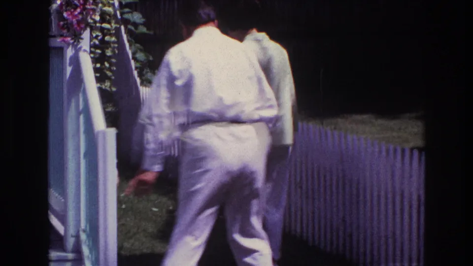 Two adults in pale 1960s-style clothing move away from white garden steps along a picket fence.