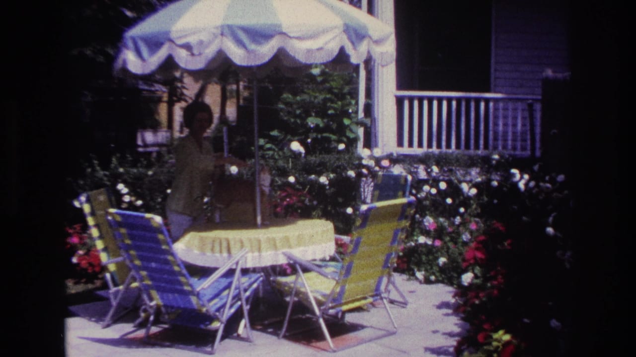 1967 Rockaway Beach home-movie frame showing a woman beside a yellow patio table, striped umbrella, flowers, greenery, and folding chairs.