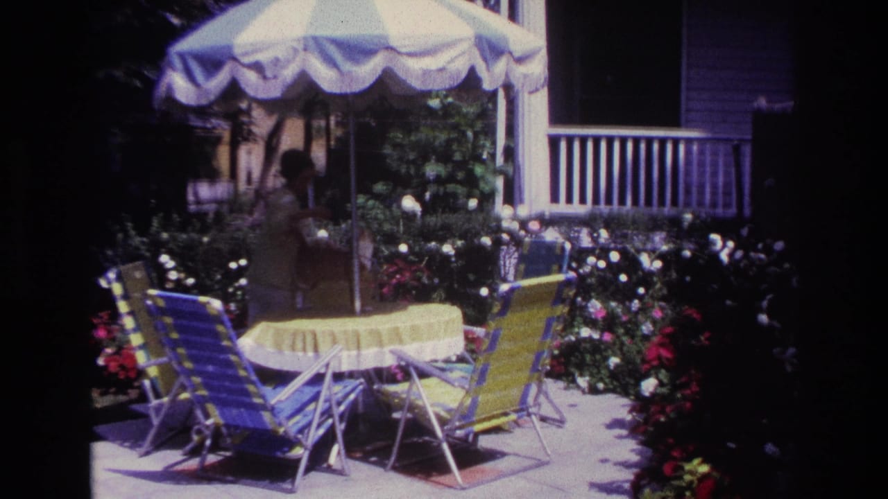 Woman reaches across the patio table under a striped umbrella with chairs and flowers around her.