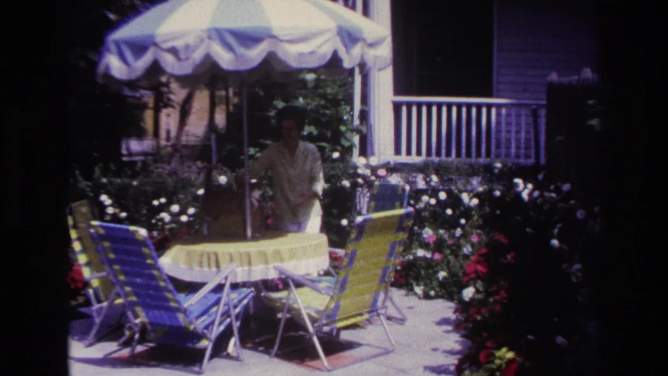 1967 Rockaway Beach frame showing a woman behind a round yellow patio table, with striped folding chairs, blooming flowers, and a house porch behind her.
