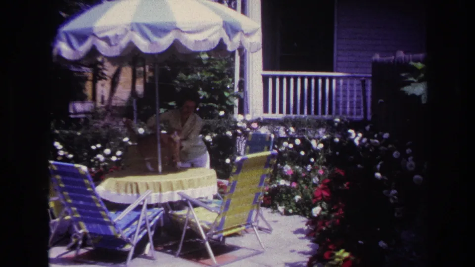 1967 Rockaway Beach home-movie frame of a woman bending over a yellow-covered patio table under a striped umbrella, likely handling a small pet.