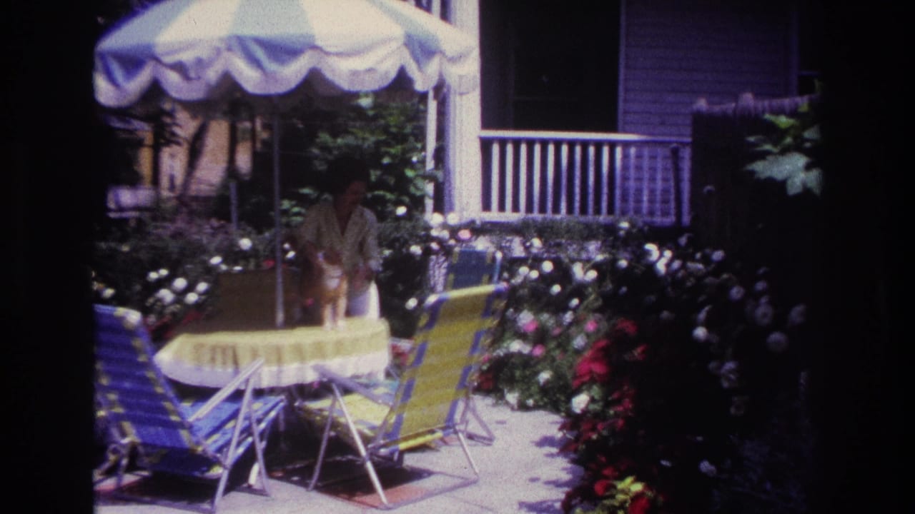1967 Rockaway Beach backyard view with a woman near a round table, striped umbrella, folding chairs, porch railing, and dense flowers.