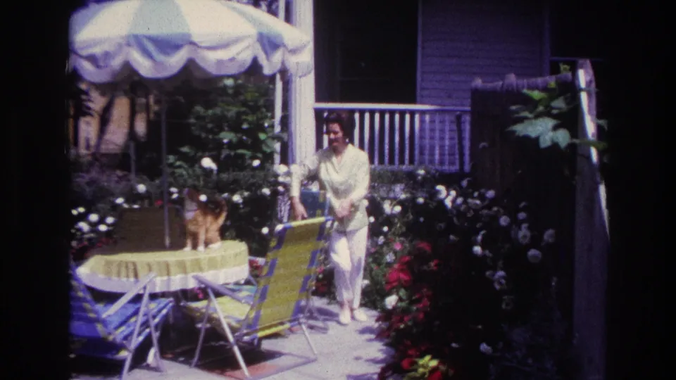 1967 Rockaway Beach home-movie frame showing a woman beside a table and a cat under a striped umbrella in a garden patio.
