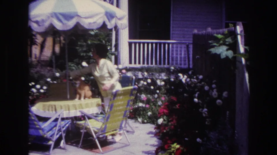 1967 Rockaway Beach home-movie frame of a woman reaching toward a cat at a yellow-covered table under a striped patio umbrella.