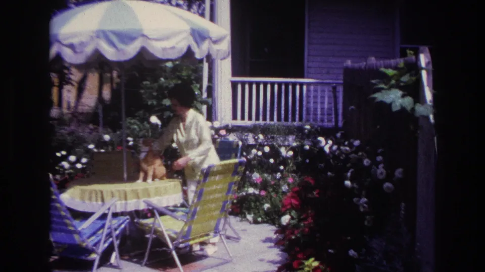 In 1967 Rockaway Beach home-movie footage, an older woman pets an orange-and-white cat on a yellow-covered patio table near flowers and chairs.