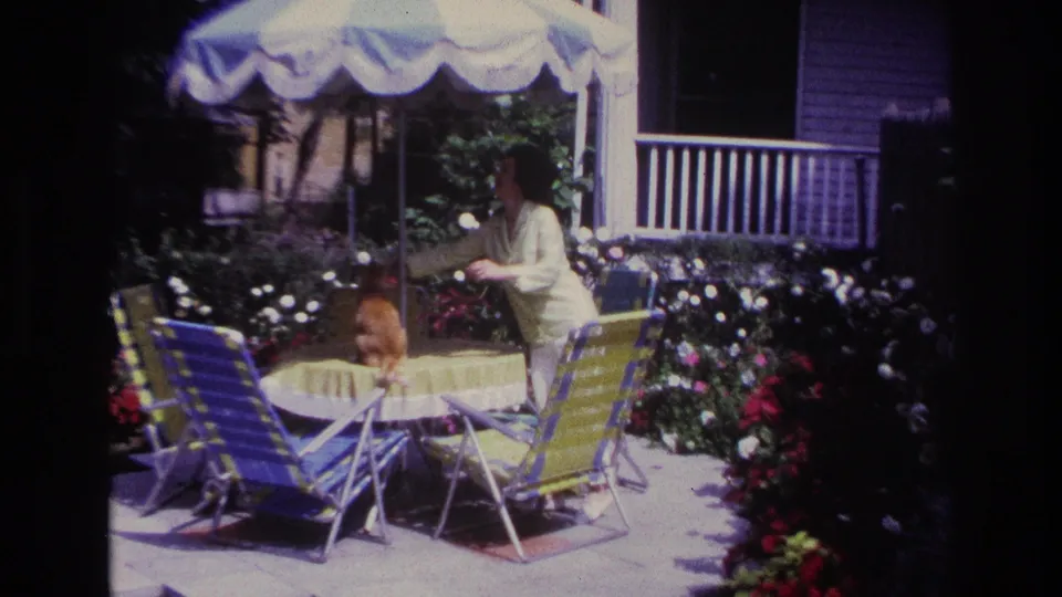 Home-movie frame from Rockaway Beach in 1967 showing a woman leaning toward an orange cat on a garden patio table.