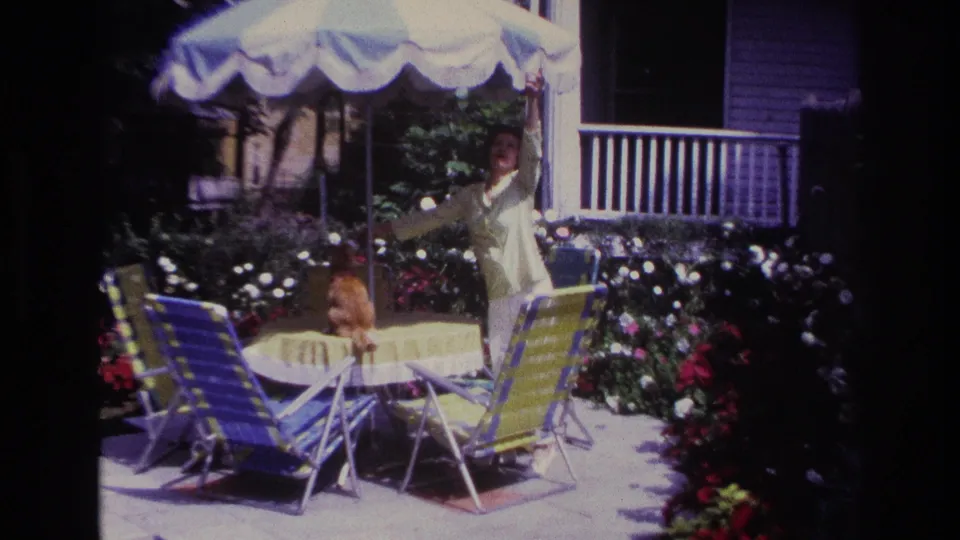 1967 Rockaway Beach home-movie frame with a woman raising her arm near an orange tabby cat on a table amid patio chairs and flowers.