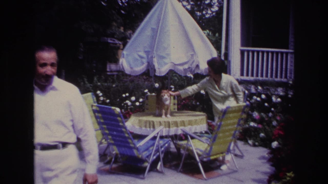 Man in white stands near a patio table as a woman pets a cat under an umbrella.