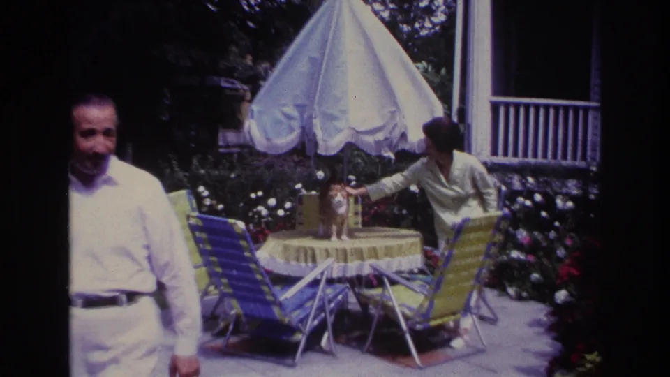 1967 Rockaway Beach home-movie frame with a man by a backyard patio table while a woman reaches toward a ginger cat under a white umbrella.