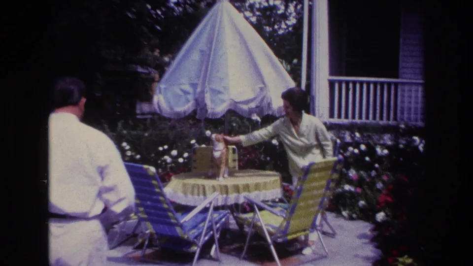 1967 Rockaway Beach 8mm frame with the man viewed from behind as the woman reaches toward the cat on the backyard patio table.