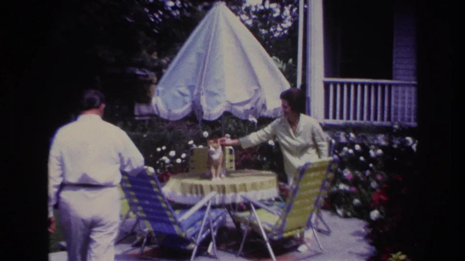 1967 Rockaway Beach home-movie frame showing the man approaching as the woman reaches to the cat beneath the white patio umbrella.