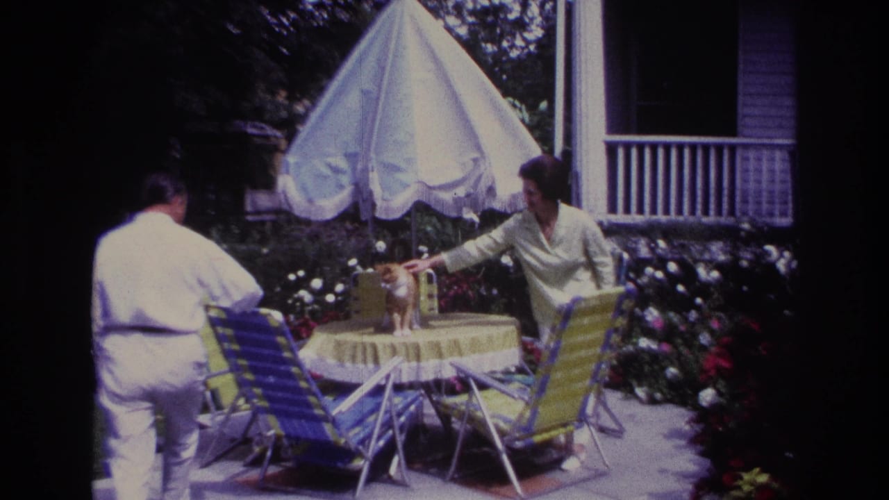1967 Rockaway Beach home-movie frame showing two adults beside a yellow patio table as a ginger cat stands near a white umbrella.