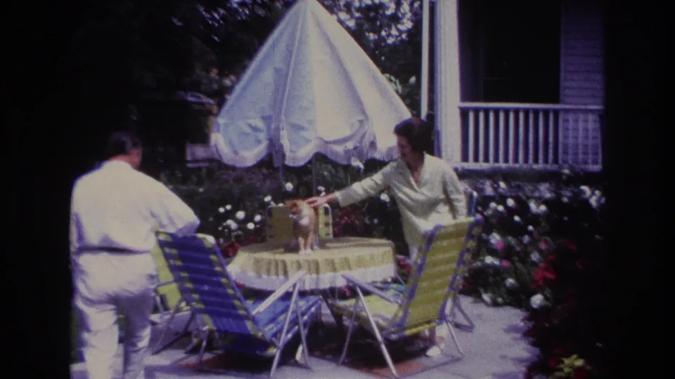 1967 Rockaway Beach home-movie frame showing two adults beside a yellow patio table as a ginger cat stands near a white umbrella.