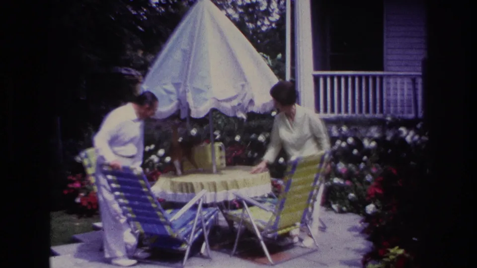 1967 Rockaway Beach patio frame showing a man bending near a striped lawn chair as a woman stands beside the table and ginger cat.