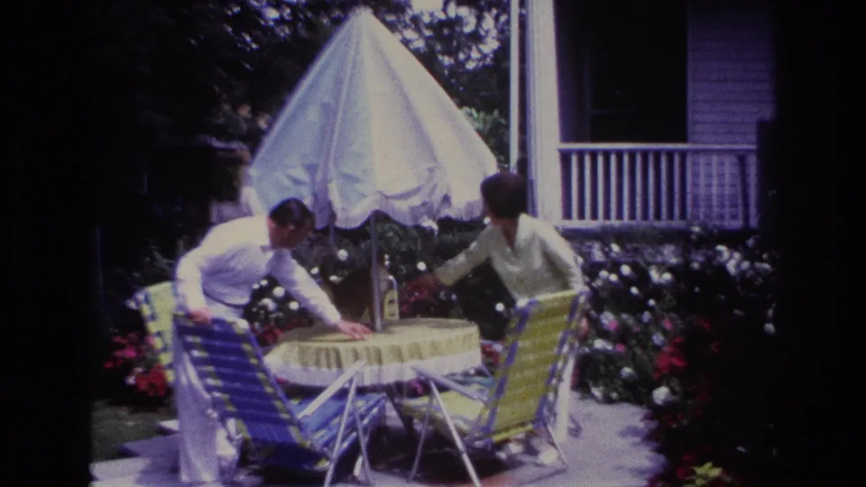 1967 Rockaway Beach home-movie frame with a man reaching over a yellow patio table as a woman and ginger cat stand in the garden.
