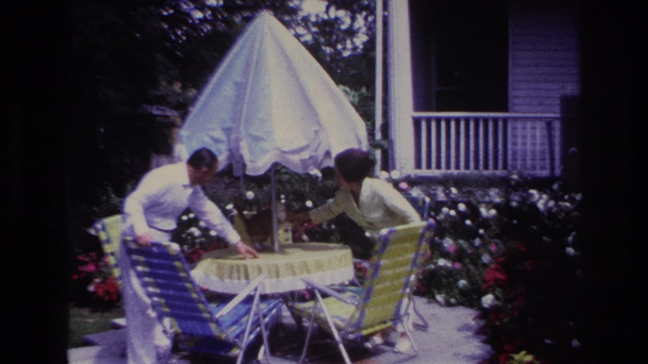 1967 Rockaway Beach home-movie frame showing a couple reaching toward a patio table beneath a white umbrella.