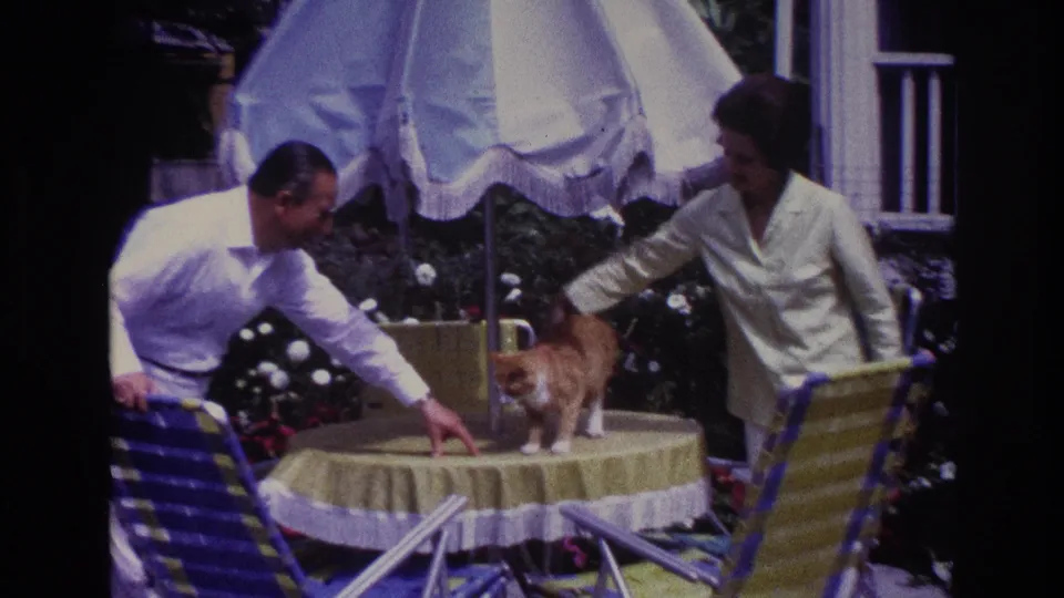 Close 1967 home-movie view of a couple beside a ginger cat on a round patio table in Rockaway Beach.