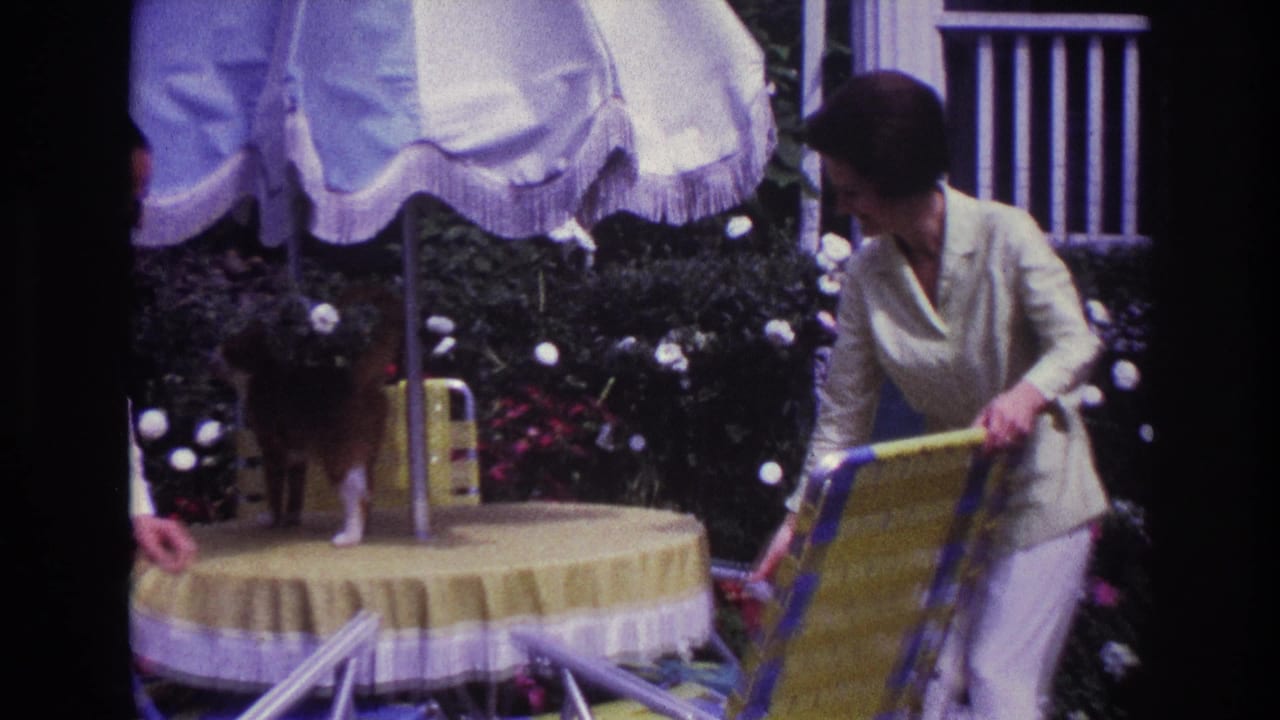 Adults setting a round patio table under a white fringed umbrella near garden flowers.